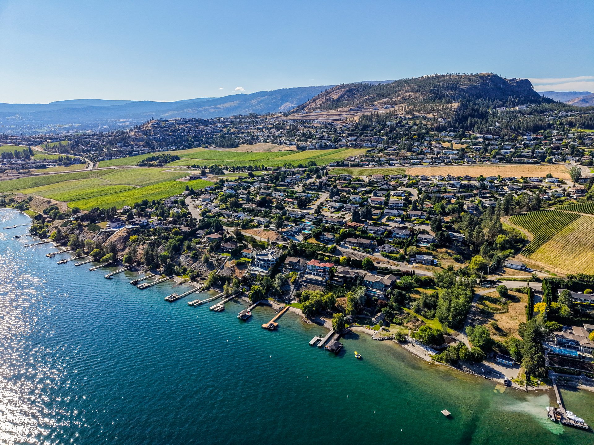 Wide aerial of Okanagan Lake shoreline with docks, vineyards and Kelowna skyline in the distance